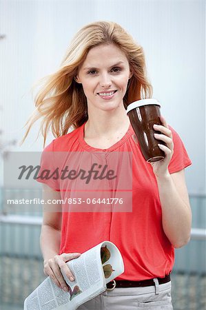 Smiling blond woman holding coffee to go and newspaper