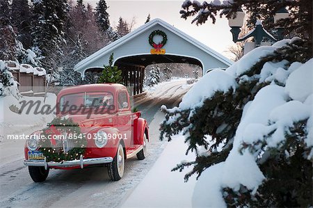 Man driving a vintage 1941 Ford pickup through a covered bridge with a Christmas wreath on the grill and a tree in the back during Winter in Southcentral, Alaska