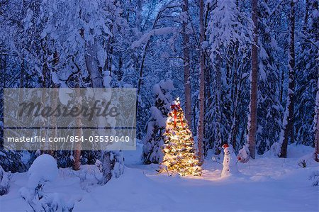 Snowman with santa hat hanging ornaments on a  Christmas tree in a snow covered birch forest in Southcentral Alaska