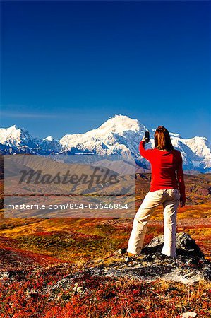 A woman hiking in Peters Hills stops to take a picture of Mt. McKinley with her cell phone, Denali State Park, Southcentral Alaska, Fall/n