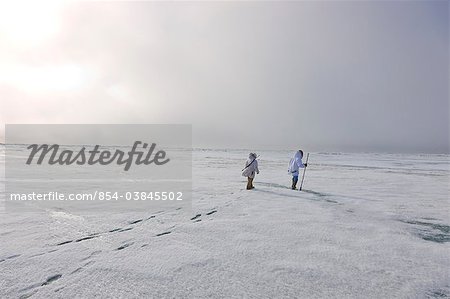 Two Inupiaq Eskimo hunters wearing their Eskimo parka's (Atigi) carry a rifle and walking stick while walking over the shore ice along the Chukchi Sea, Barrow, Arctic Alaska, Summer