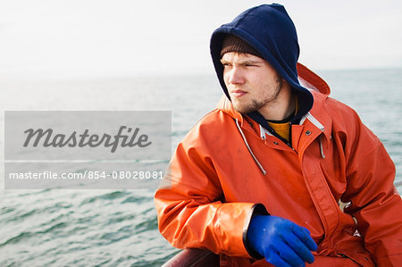 A Rain Gear Clad Deckhand Takes A Break And Sits On The Rail Looking Out At The Net While Salmon Fishing On The Copper River Flats; Cordova Alaska United States Of America