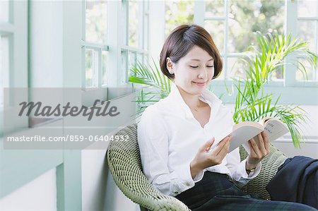 Japanese woman in a white shirt reading a book on a wicker chair