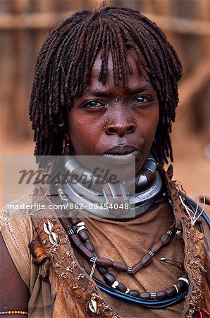 A Hamar woman in the village square of Dimeka. Married women wear two heavy steel necklaces. This woman wears an extra necklace with steel a steel phallic symbol which identifies her as a first wife. She wears her hair long in a braided fringe matted with animal fat and ochre.