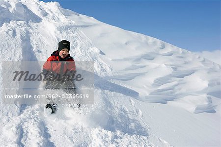 Norway,Tromso,Lyngen Alps. Young boy sits on a tobogganing seat to slide down a steep snow bank in bright winter sunshine