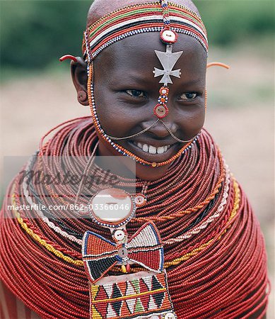 Samburu girls are given strings of beads by their fathers when they are still young. As soon as they are old enough to have lovers from the warrior age-set,they regularly receive gifts from them. Over a period of years,their necklaces can smother them up to their necks. The metal cross-like ornament hanging from the girl's headband has no religious significance.