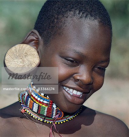 A young Maasai girl wearing a wooden plug in her pierced ear to elongate the earlobe. It has been a tradition of the Maasai for both men and women to pierce their ears and elongate their lobes for decorative purposes. Her two lower incisors have been removed - a common practice that may have resulted from an outbreak of lockjaw a long time ago.