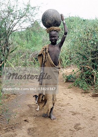A Mursi girl, accompanied by her dog, carries a large clay pot to collect water from the Omo River. Her earlobes are already pierced and extended, and decorated with round clay discs.She is dressed in skins, attractively decorated with thin stripes.The culture, social organisation, customs and values of the people have changed little.