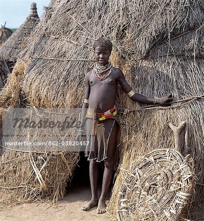 A Karo girl in traditional attire stands outside her familys home.The door of the low entrance to the house is propped against its thatched wall.Most girls pierce a hole below the lower lip in which they place a thin piece of metal or a nail for decoration.The Karo are a small tribe living in three main villages along the lower reaches of the Omo River in southwest Ethiopia.