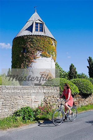 France, Charente Maritime, Ile de Re.  A tourist cycles past an old windmill now converted into a house on the outskirts of Les Portes. MR