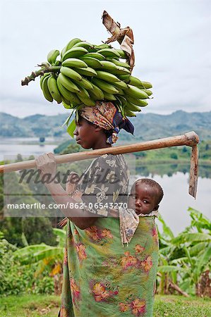 A Ugandan woman returns from her farm with bananas balanced on her head, her child slung comfortably on her back, Uganda, Africa