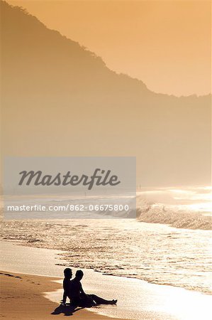 South America, Rio de Janeiro, Rio de Janeiro city, a gay couple silhouetted against in golden light, sit on the sand on Copacabana Beach with Copacabana and the Morro do Leme hill in the background