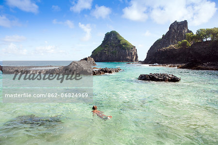 South America, Brazil, Pernambuco, Fernando de Noronha Island, a young woman snorkelling at the Bay of Pigs with the Two Brothers rocks behind MR
