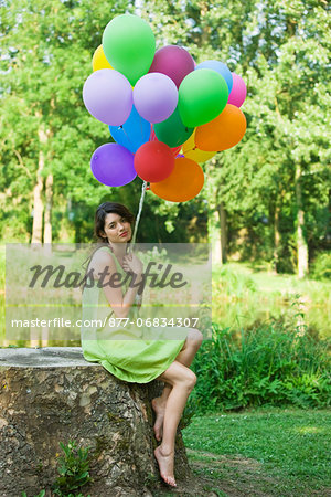 Young woman holding bunch of balloons, sitting on tree stump