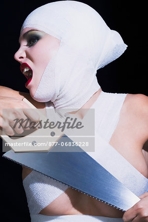 Portrait of a young woman shouting, holding handsaw, bandages on head and breast