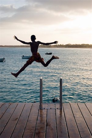 A boy playing a game jumps into the air from a wooden jetty inthe Cape Verde islands.He has his arms and legs spread wide and is in silhouette against the sky. Foto de stock - Super Valor sin royalties y Suscripción, Código: 400-04018376