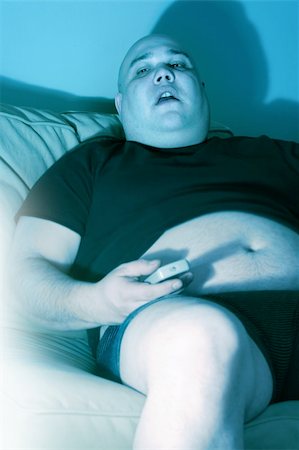 slob on couch - Lazy overweight male sitting on a couch watching television.  Harsh blue lighting from television with slow shutter speed to create TV watching atmosphere.  Selective focus on the eyes. Stock Photo - Budget Royalty-Free & Subscription, Code: 400-04011189