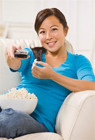 A beautiful young asian woman sitting on a couch. She is holding a bowl of popcorn and is using a remote. She is smiling directly at the camera. Vertically framed shot. Stock Photo - Budget Royalty-Free & Subscription, Code: 400-04119208