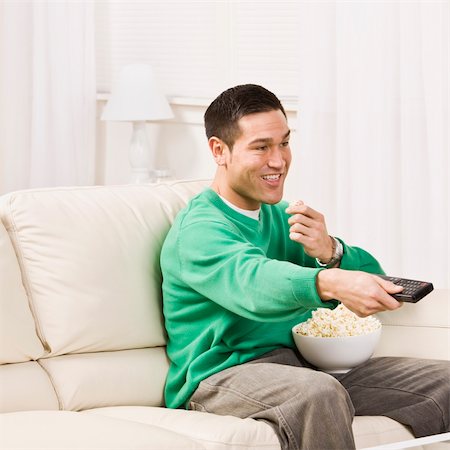 A man seated on a couch and using a remote control for T.V. He is eating popcorn and smiling. Square framed photo. Stock Photo - Budget Royalty-Free & Subscription, Code: 400-04120526