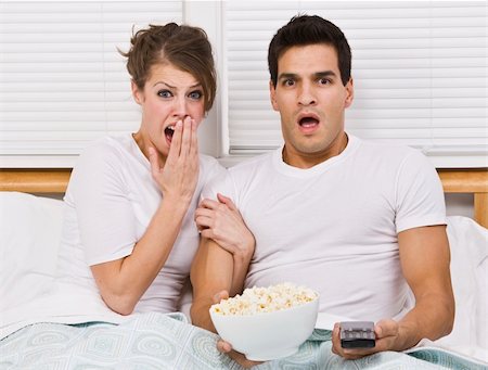 A young, attractive couple is sitting together in bed and watching TV.  They look shocked or scared, and are looking away from the camera.  Horizontally framed shot. Stock Photo - Budget Royalty-Free & Subscription, Code: 400-04120679
