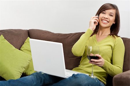 A beautiful young oriental woman with a wonderful toothy smile drinking redwine, chatting on her cell phone and using a laptop on her settee Stock Photo - Budget Royalty-Free & Subscription, Code: 400-04132589