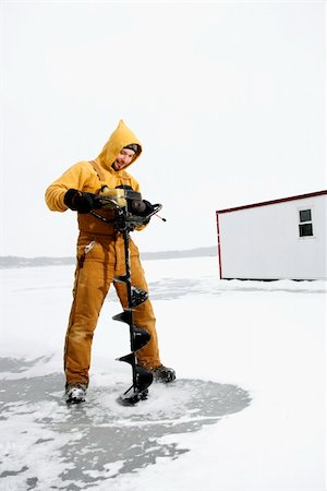 pesca en el hielo - Young man in yellow snow gear smiles as he drills a hole in the ice for fishing. Vertical shot. Foto de stock - Super Valor sin royalties y Suscripción, Código: 400-04169298
