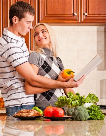 simsearch:400-04162443,k - Young woman looks back affectionately at a young man as he reads a recipe book. A kitchen counter holds a variety of fresh vegetables in the foreground. Vertical shot. Fotografie stock - Microstock e Abbonamento, Codice: 400-04162449