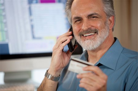 A businessman holding up a credit card and making a payment on his cellphone with a computer monitor in the background. Horizontal shot. Stock Photo - Budget Royalty-Free & Subscription, Code: 400-04172658