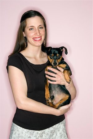 simsearch:400-04446187,k - Smiling Caucasian woman holding Miniature Pinscher dog standing against pink background. Photographie de stock - Aubaine LD & Abonnement, Code: 400-04446310