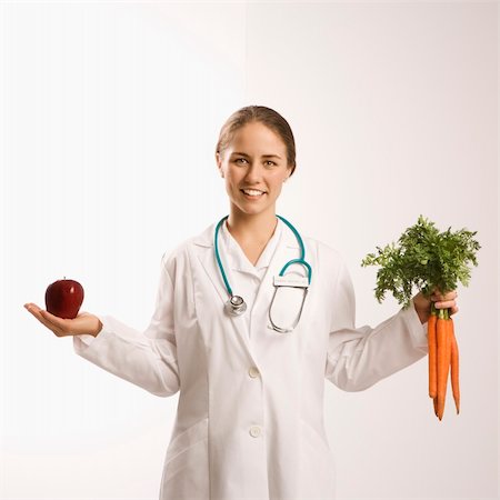 simsearch:400-04956215,k - Portrait of Caucasian mid-adult female doctor holding red apple and bunch of carrots smiling and looking at viewer. Photographie de stock - Aubaine LD & Abonnement, Code: 400-04452069