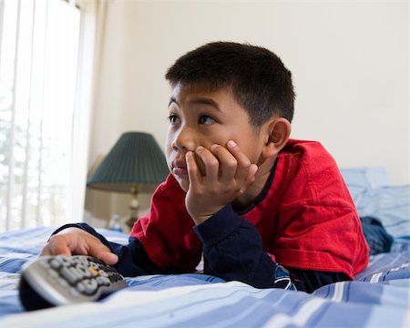 Young asian boy laying down on his stomach on the bed while looking engrossed in the television holding the remote control Stock Photo - Budget Royalty-Free & Subscription, Code: 400-04503258