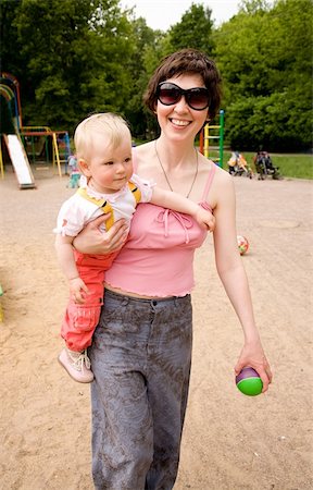 simsearch:400-06073989,k - Beautiful harmonous mum with dark hair in sun glasses carries a little daughter and a green ball on a children's playground Photographie de stock - Aubaine LD & Abonnement, Code: 400-04570609