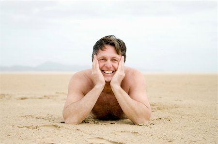 A happy smiling forties man is laying on his stomache on a beautiful empty sandy beach on the island of fuerteventura in the canary islands. Foto de stock - Super Valor sin royalties y Suscripción, Código: 400-04618488