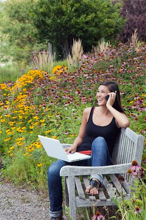 Cute young woman laughs while talking on the phone and working on a laptop. She is seated on a park bench amid blooming flowers and trees. Vertical shot. Stock Photo - Budget Royalty-Free & Subscription, Code: 400-04714202