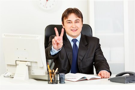 diretor de escola - Smiling modern business man sitting at office desk and showing victory gesture Foto de stock - Royalty-Free Super Valor e Assinatura, Número: 400-04794636