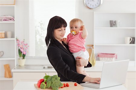 simsearch:400-04712944,k - Beautiful brunette woman in suit holding her baby in her arms while working with her laptop in the kitchen Stock Photo - Budget Royalty-Free & Subscription, Code: 400-04884721