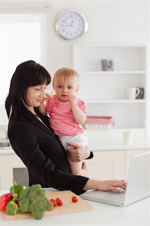 simsearch:400-04712944,k - Attractive brunette woman in suit holding her baby in her arms while working with her laptop in the kitchen Stock Photo - Budget Royalty-Free & Subscription, Code: 400-04884722