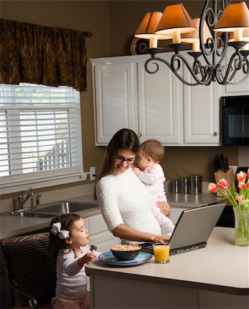 Caucasian mother holding baby  and typing on laptop computer with girl eating breakfast in kitchen. Stock Photo - Budget Royalty-Free & Subscription, Code: 400-05038976