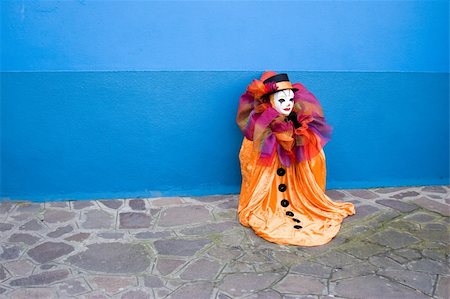 Small orange Clown in white mask, and top hat sitting in front of the blue wall - alone Foto de stock - Royalty-Free Super Valor e Assinatura, Número: 400-05031263