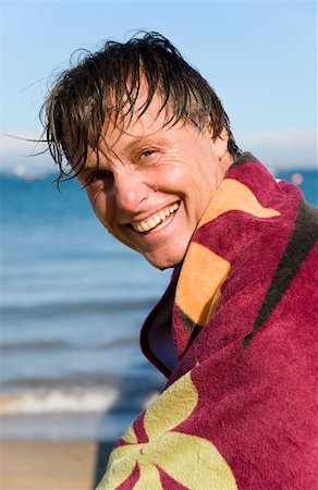 A handsome smiling forties man is sat on a beach with a towel wrapped around him to keep warm. Foto de stock - Super Valor sin royalties y Suscripción, Código: 400-05149578