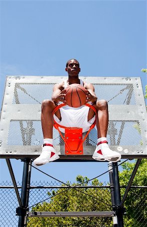 Handsome sporty African-American male basketball player dressed in white and holding his ball outdoor on a summer day in a basketball court while sitting in the hoop. Stock Photo - Budget Royalty-Free & Subscription, Code: 400-05155908