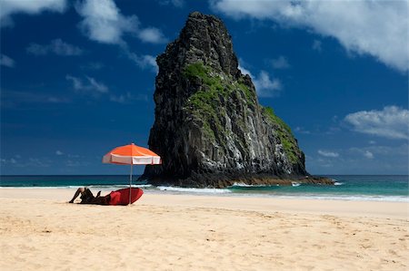 simsearch:400-03985677,k - A young surfer is laying under an umbrella waiting for waves. The deserted beach is shot against deep blue skies with few clouds and a huge rock in the middle. The picture is taken on Fernando de Noronha Island, Brazil. Foto de stock - Super Valor sin royalties y Suscripción, Código: 400-05232181