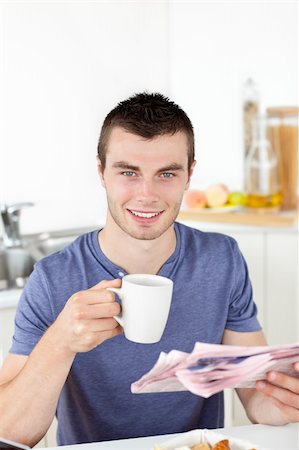 simsearch:400-04209411,k - Positive young man holding a cup and a newspaper smiling at the camera sitting in the kitchen Fotografie stock - Microstock e Abbonamento, Codice: 400-05249130