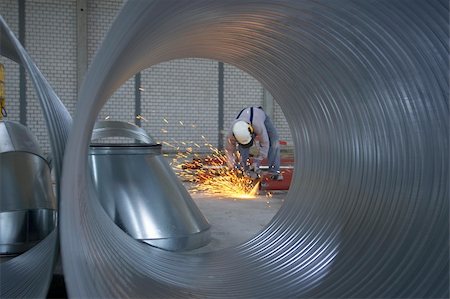 Pipe view towards an industrial worker in safety workwear and white helmet cutting iron pipes for a ventilation system made to specification using an angle grinder producing sparks in a spaceous factory building. Corrugated aluminum pipes with a big diameter in front of the scenery. Foto de stock - Super Valor sin royalties y Suscripción, Código: 400-05284829