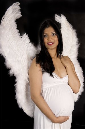 A beautiful pregnant Indian woman in white dress and angel wings smiling on black backdrop Stockbilder - Microstock & Abonnement, Bildnummer: 400-05302904