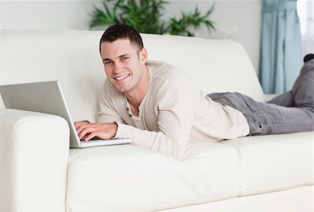 simsearch:400-05715585,k - Smiling man lying on a sofa with a laptop while looking at the camera Foto de stock - Super Valor sin royalties y Suscripción, Código: 400-05715659