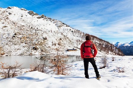 One man young male hiker stands in front of a alpine lake looking to the snowy landscape, rear view. Red jacket, black pants. Large copy-space at top-right. Buffet Lake, Champorcher, Val d'aosta, Italy, Europe. Stock Photo - Budget Royalty-Free & Subscription, Code: 400-05730784
