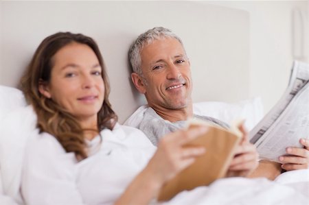 simsearch:400-05749442,k - Happy woman reading a book while her husband is reading the news in their bedroom Photographie de stock - Aubaine LD & Abonnement, Code: 400-05749451