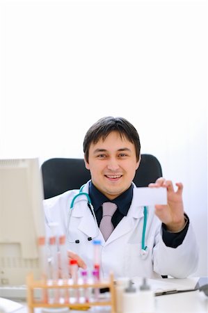 simsearch:400-07099889,k - Smiling doctor sitting at office table and holding blank business card Stock Photo - Budget Royalty-Free & Subscription, Code: 400-05883650