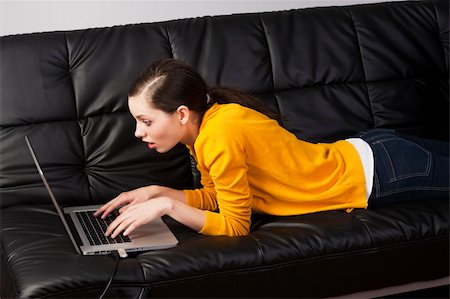 simsearch:400-04999709,k - very young girl in yellow shirt laying an a black sofa and working on laptop computer, she's liyng on the the sofa, watches the laptop's display with expression of surprise Photographie de stock - Aubaine LD & Abonnement, Code: 400-05890359